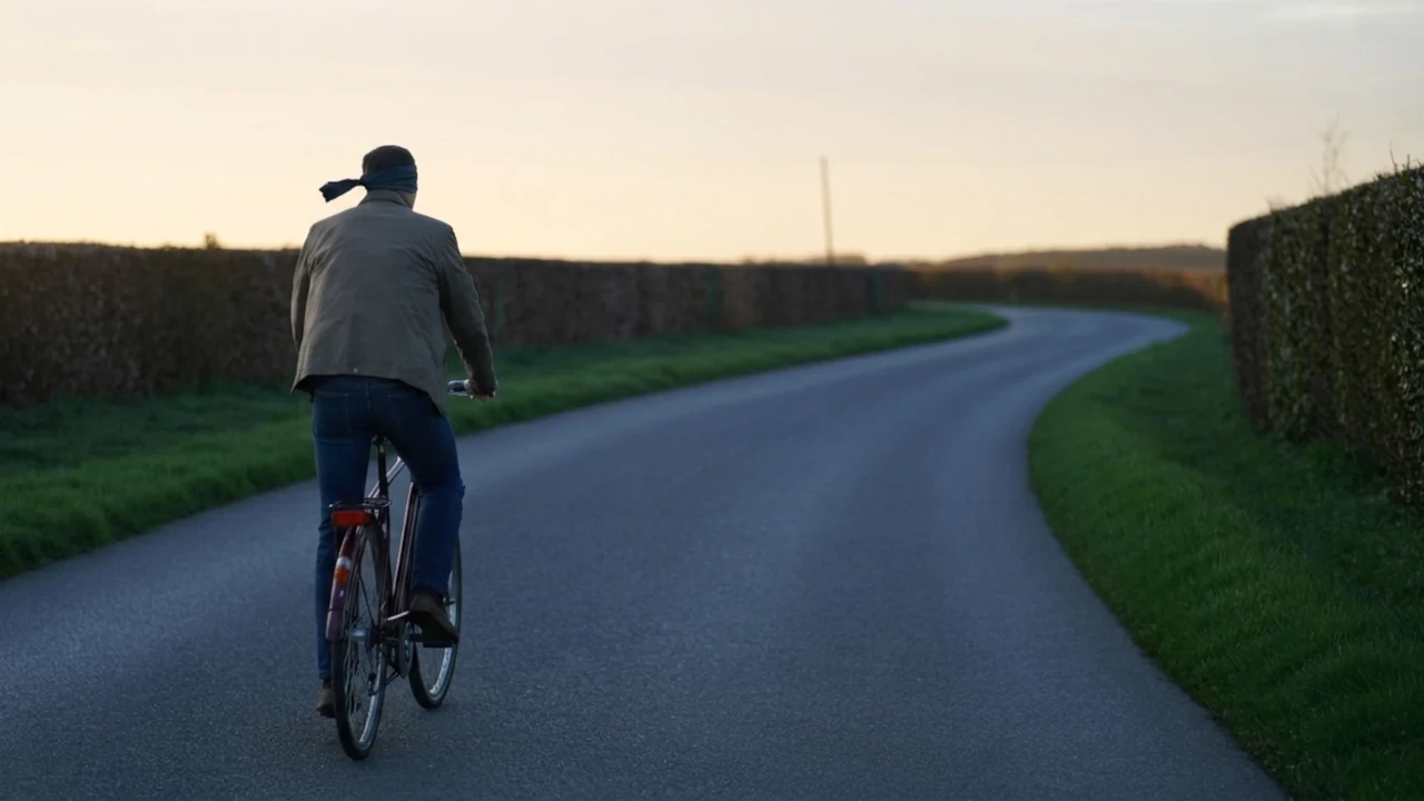 Blindfolded man cycling away down a narrow hedge-lined lane at dusk, the road curving out of sight, moving forward without a clear strategy.